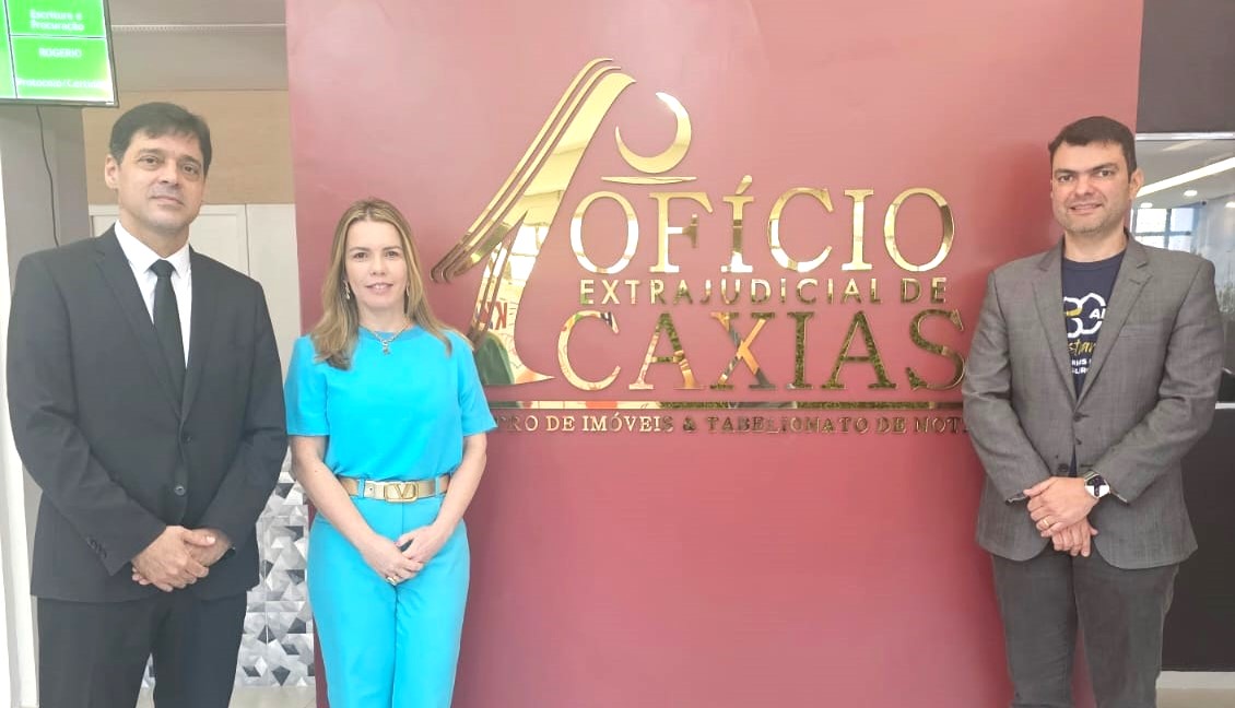 A foto mostra dois homens e uma mulher em pé em frente a uma parede vermelha com letras douradas fundo com nome "1º OFÍCIO EXTRAJUDICIAL DE CAXIAS".  À Esquerda, homem pardo, de terno preto, ao lado de mulher branca, cabelos louros, de calça e blusa azuis e à direita, homem branco, de terno cinza e camiseta preta com letras brancas.