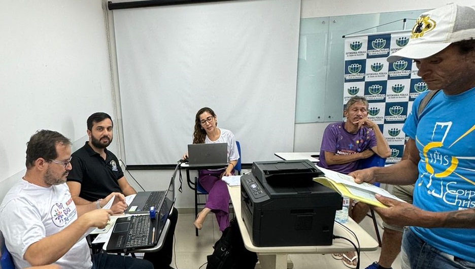 Foto horizontal, colorida. Duas mulheres e dois homens esto sentados em uma sala de paredes claras.  direita, DE P, um homem negro, de camiseta azul com estampa colorida e bon branco, segura documentos. Ao fundo, um quadro branco na parede e vrios logotipos coloridos.