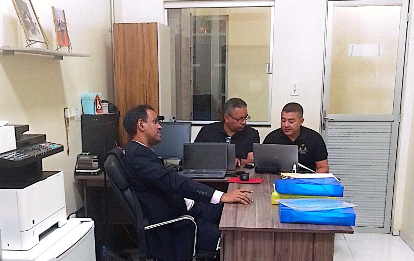 Foto horizontal, colorida, de três homens em sala de trabalho, junto a mesas e computadores. Os homens vestem trajes formais, de cores escuras e estão sentados. Ao fundo, parede com janelas de vidro.