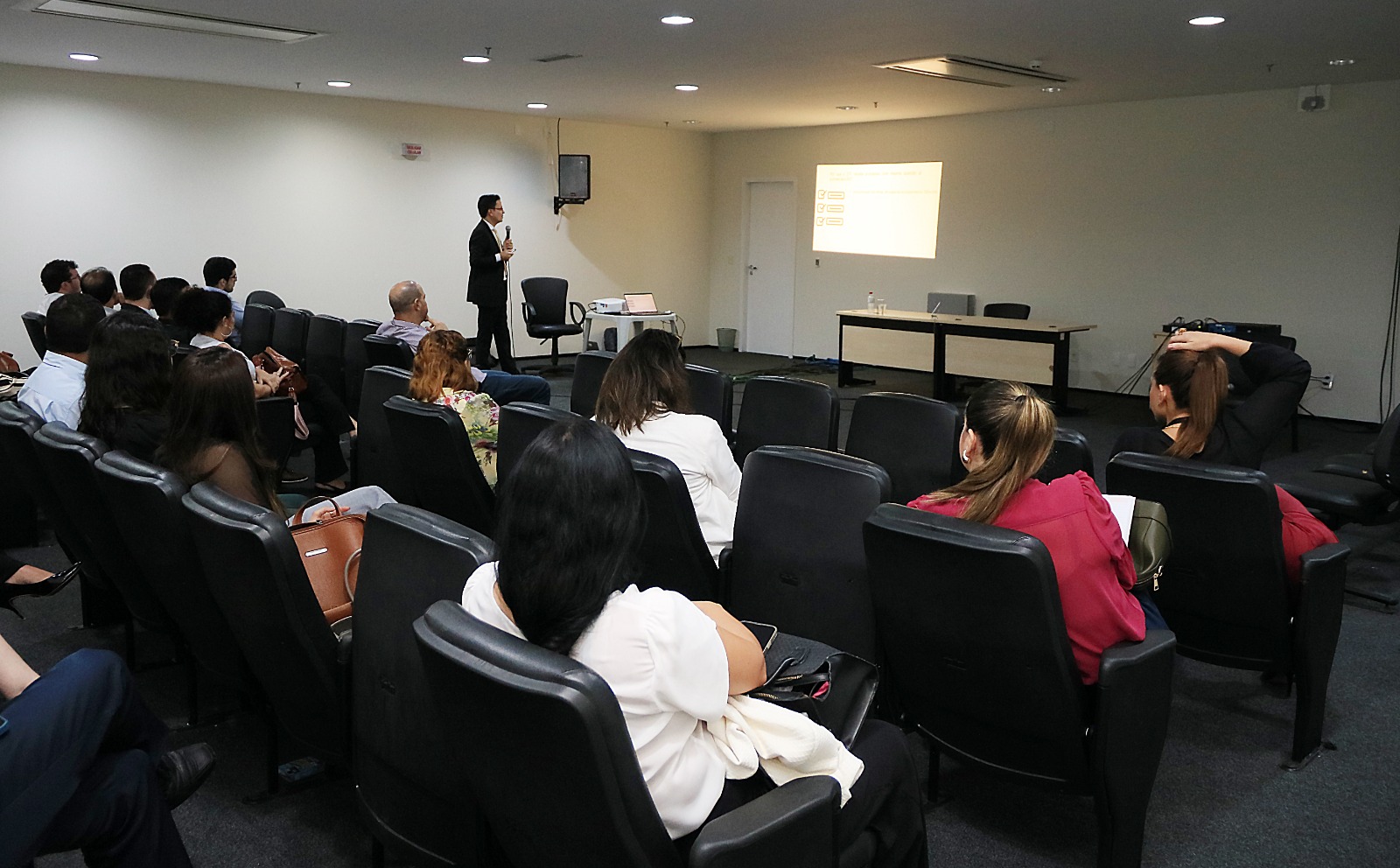 A imagem mostra um grupo de pessoas sentadas em uma sala de conferências, participando de uma apresentação. O apresentador está de pé perto de uma tela de projetor, segurando um microfone, enquanto parece explicar o conteúdo exibido na tela. Os participantes estão atentos à apresentação.

A sala possui várias fileiras de cadeiras, uma mesa com alguns itens sobre ela e um laptop conectado ao projetor. O slide projetado contém texto, mas não é completamente legível na imagem. O ambiente sugere um contexto profissional ou educacional, voltado para discussões, aprendizado ou compartilhamento de informações.
