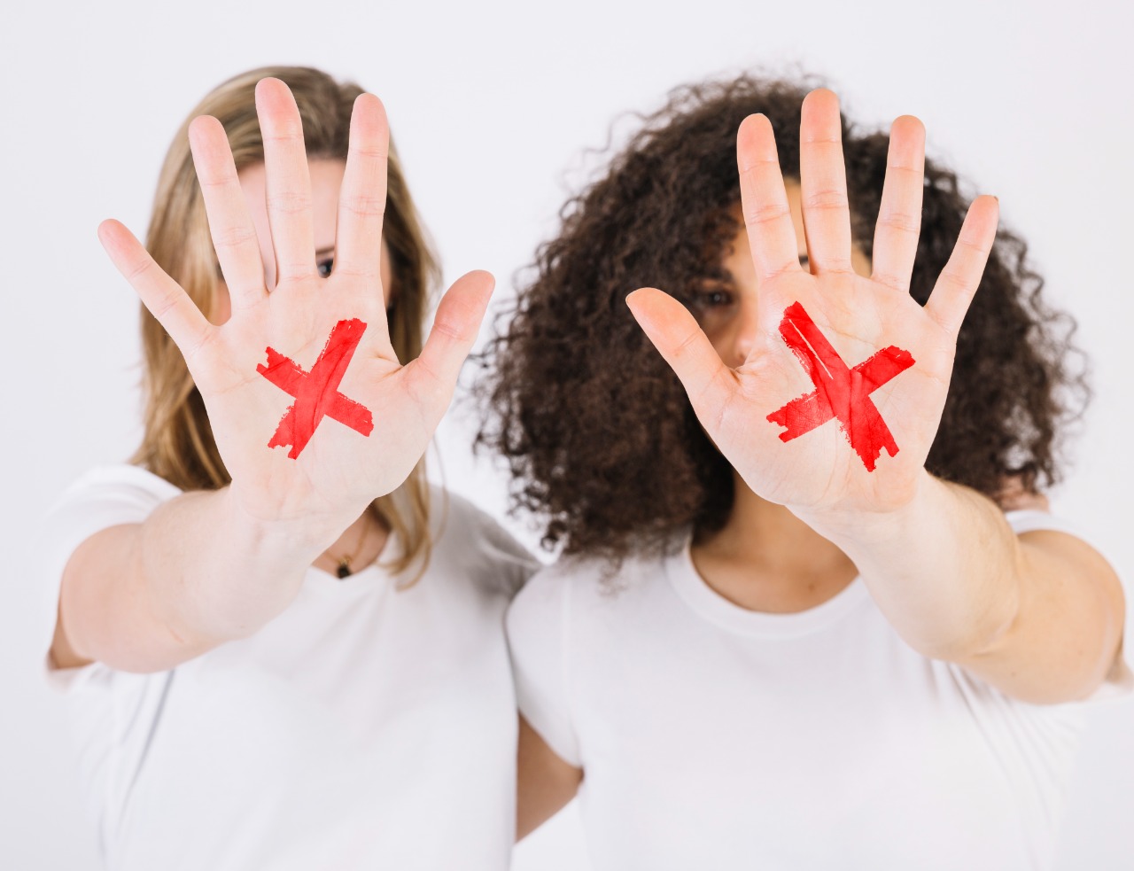 Imagem colorida. Na foto aparece duas mulheres,, de frente, com os braços estendidos aparecendo uma das palmas das mãos de cada uma, nelas há um "x" vermelho.