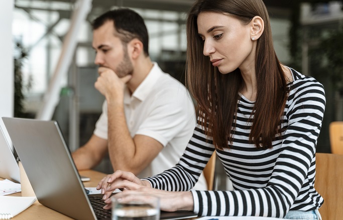 Foto colorida ilustrativa. Uma mulher em primeiro plano digita em notebook. Um homem trabalha ao fundo em computador.">