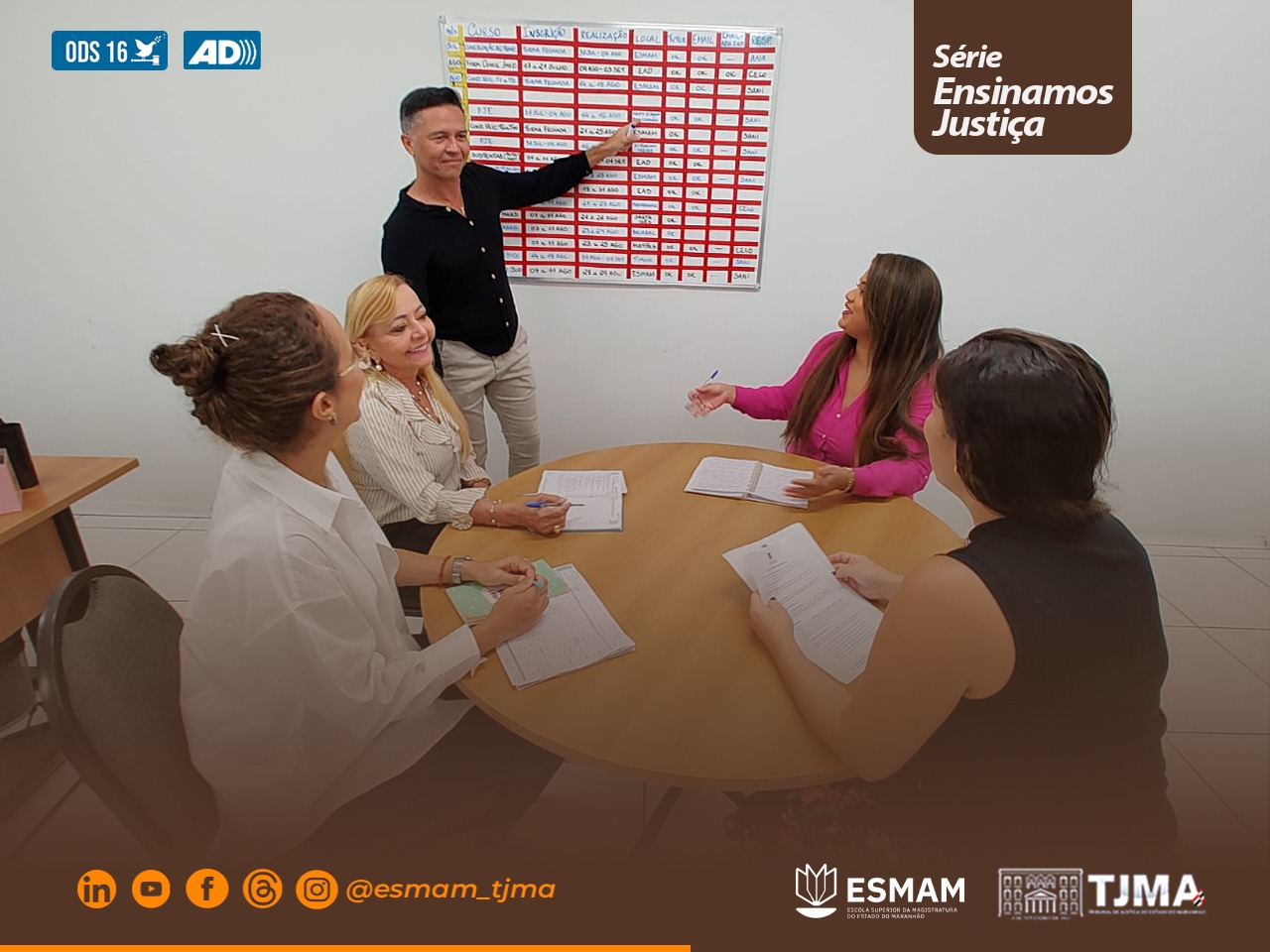 Foto horizontal. Equipe da divisão de treinamento da esmam, responsável pelas ações de formação do judiciário do maranhão, reunida. um dos integrantes está em pé, homem de estatura média, usa calça jeans clara e camisa preta. ele aponta para um quadro branco, dividido em linhas e colunas, com anotações das comarcas, durante ação de planejamento dos cursos ofertada pela divisão. as demais integrantes estão sentadas, acompanhando a explicação.