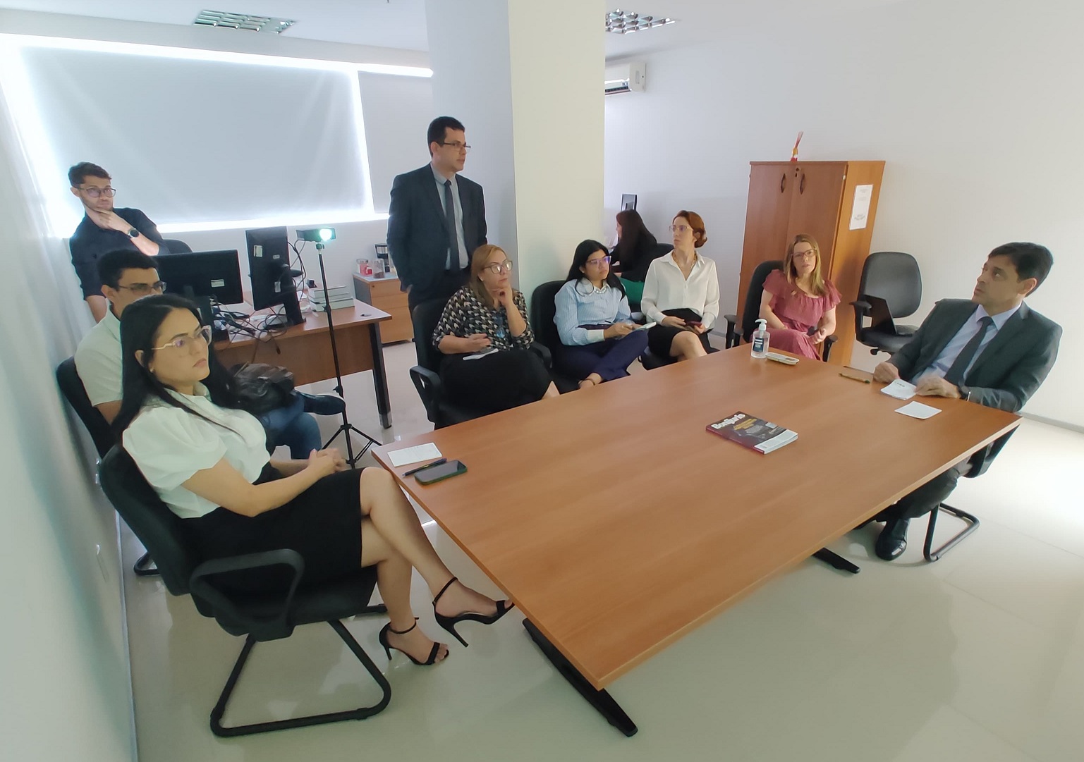 PRIMEIRA FOTO RETANGULAR DE UMA SEQUENCIA DE QUATRO IMAGENS. MOSTRA VÁRIAS PESSOAS REUNIDAS EM TORNO DE UMA MESA DE REUNIÃO. ALGUMAS INTERAGEM ENTRE SI E OUTRAS OLHAM PARA DIREÇÃO ONDE É FEITA A PROJEÇÃO DE UMA APRESENTAÇÃO.