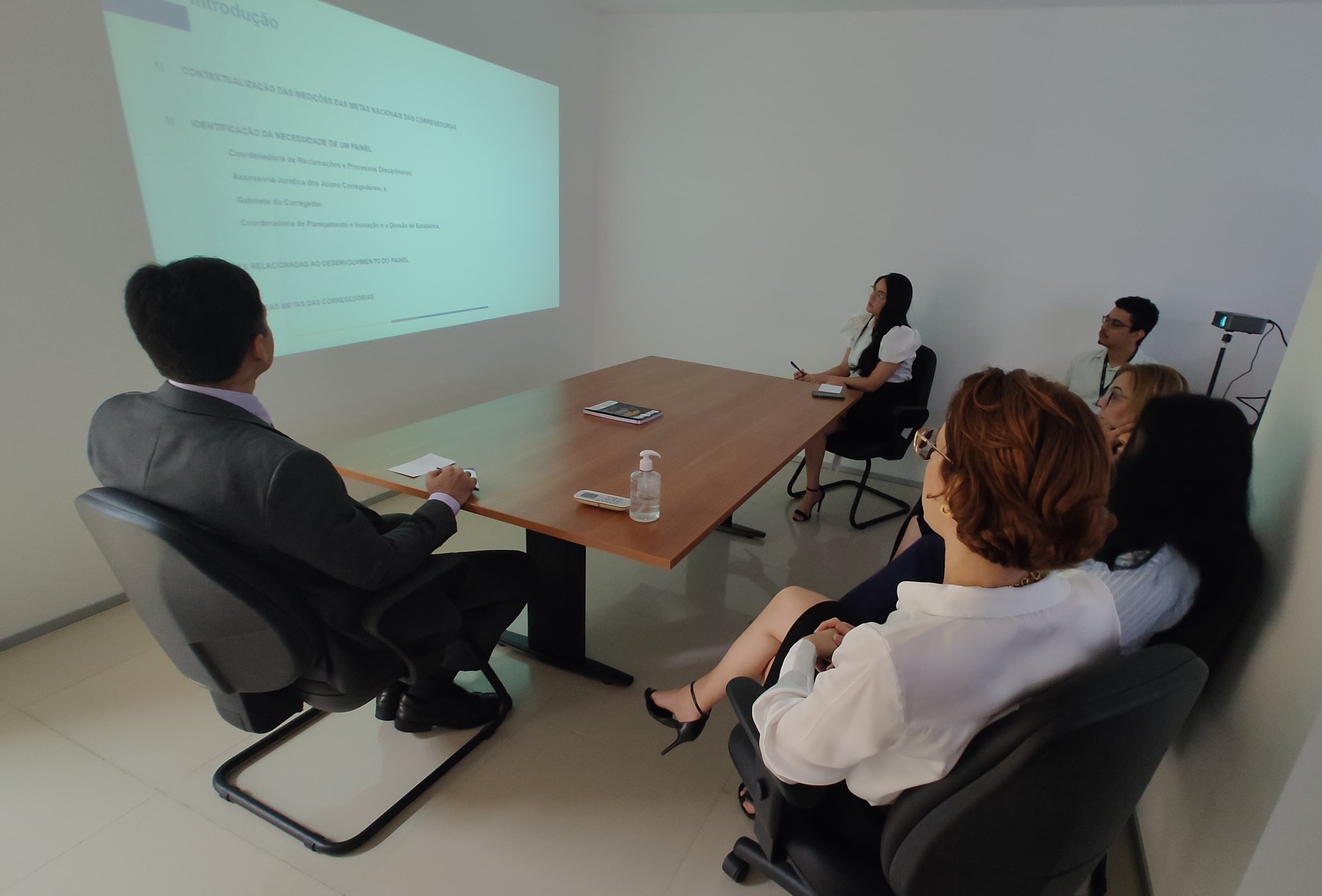 TERCEIRA FOTO RETANGULAR DE UMA SEQUENCIA DE QUATRO IMAGENS. MOSTRA VÁRIAS PESSOAS REUNIDAS EM TORNO DE UMA MESA DE REUNIÃO. ALGUMAS INTERAGEM ENTRE SI E OUTRAS OLHAM PARA DIREÇÃO ONDE É FEITA A PROJEÇÃO DE UMA APRESENTAÇÃO.