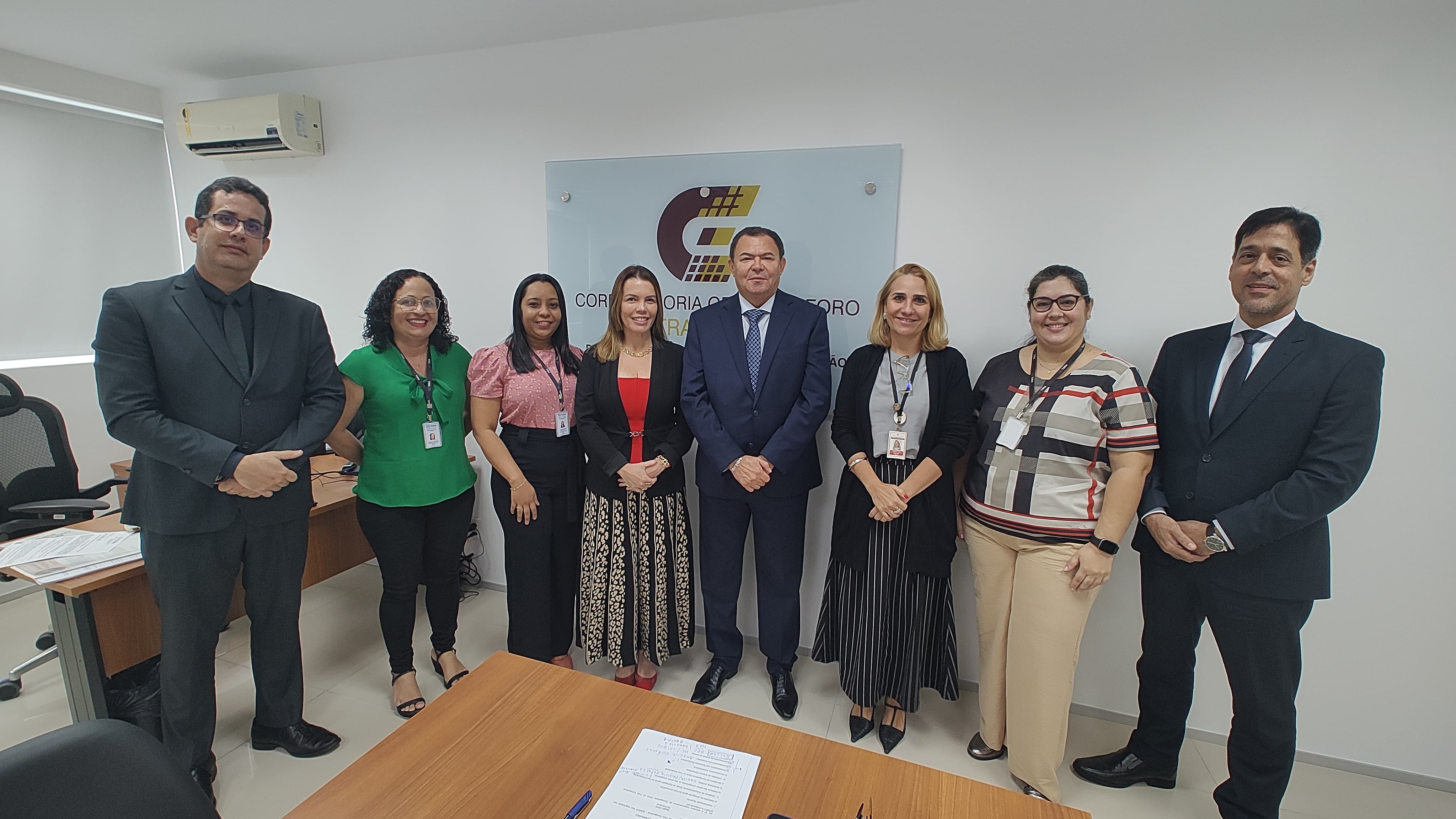 Imagem da reunião com a Diretoria de Auditoria Interna do TJMA, realizada na sede da COGEX. Mostra integrantes da Diretoria e da corregedoria extrajudicial após a reunião. Estão todas e todos em pé, lado a lado, posando para foto. Há uma mesa de trabalho marrom, cadeiras pretas e paredes brancas. A reunião foi para tratar da continuidade da consultoria para desenvolver o planejamento estratégico da COGEX.