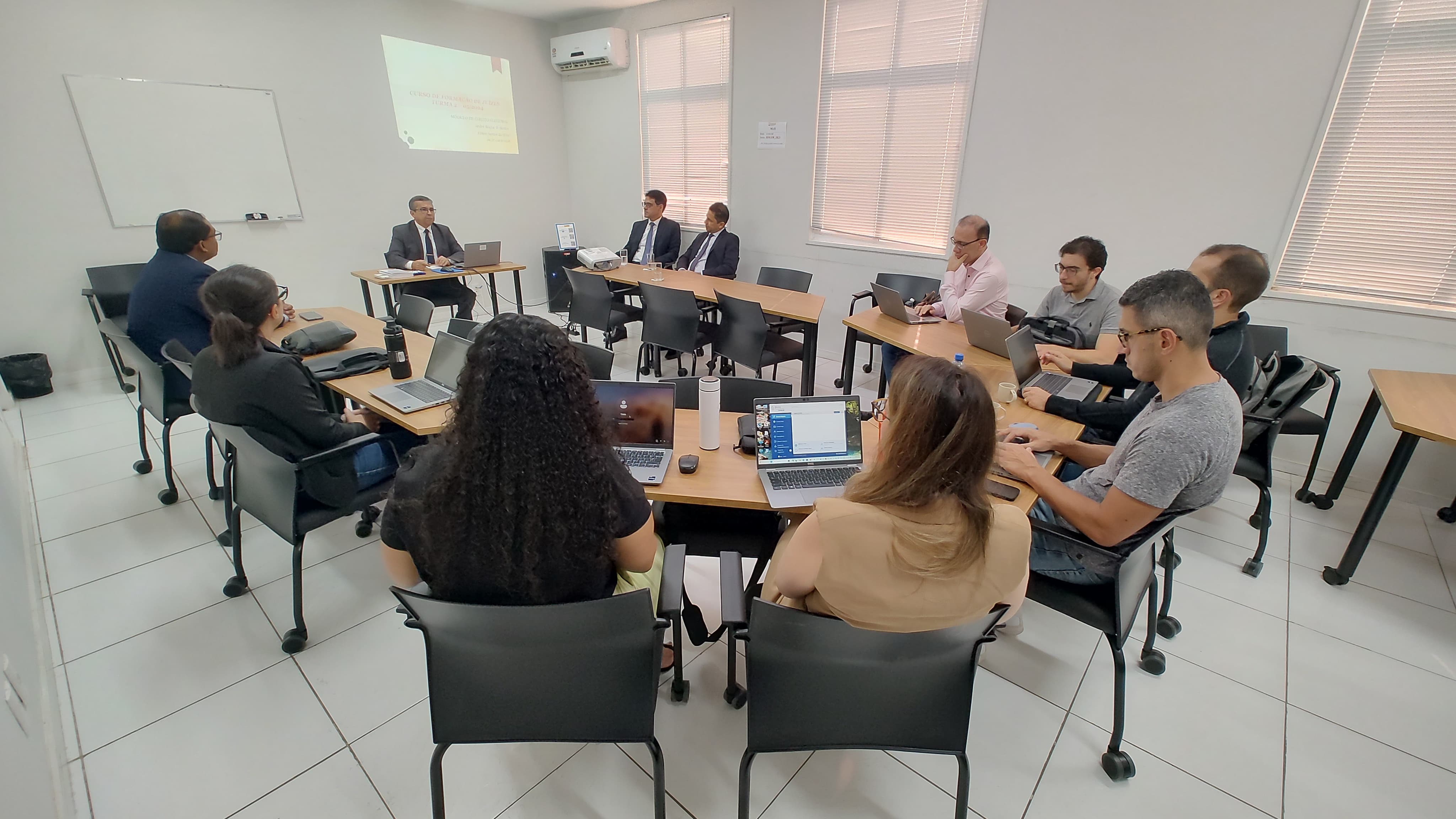 Fotografia horizontal. mostra sala de aula da esmam, que possui piso, teto e paredes brancas. a perspectiva é do fundo para a frente da sala. mostra alunas e alunos estão sentados e dispostos em formato circular e ouvem atentamente a exposição feita pelo desembargador josé gonçalo, presidente do tribunal regional eleitoral.