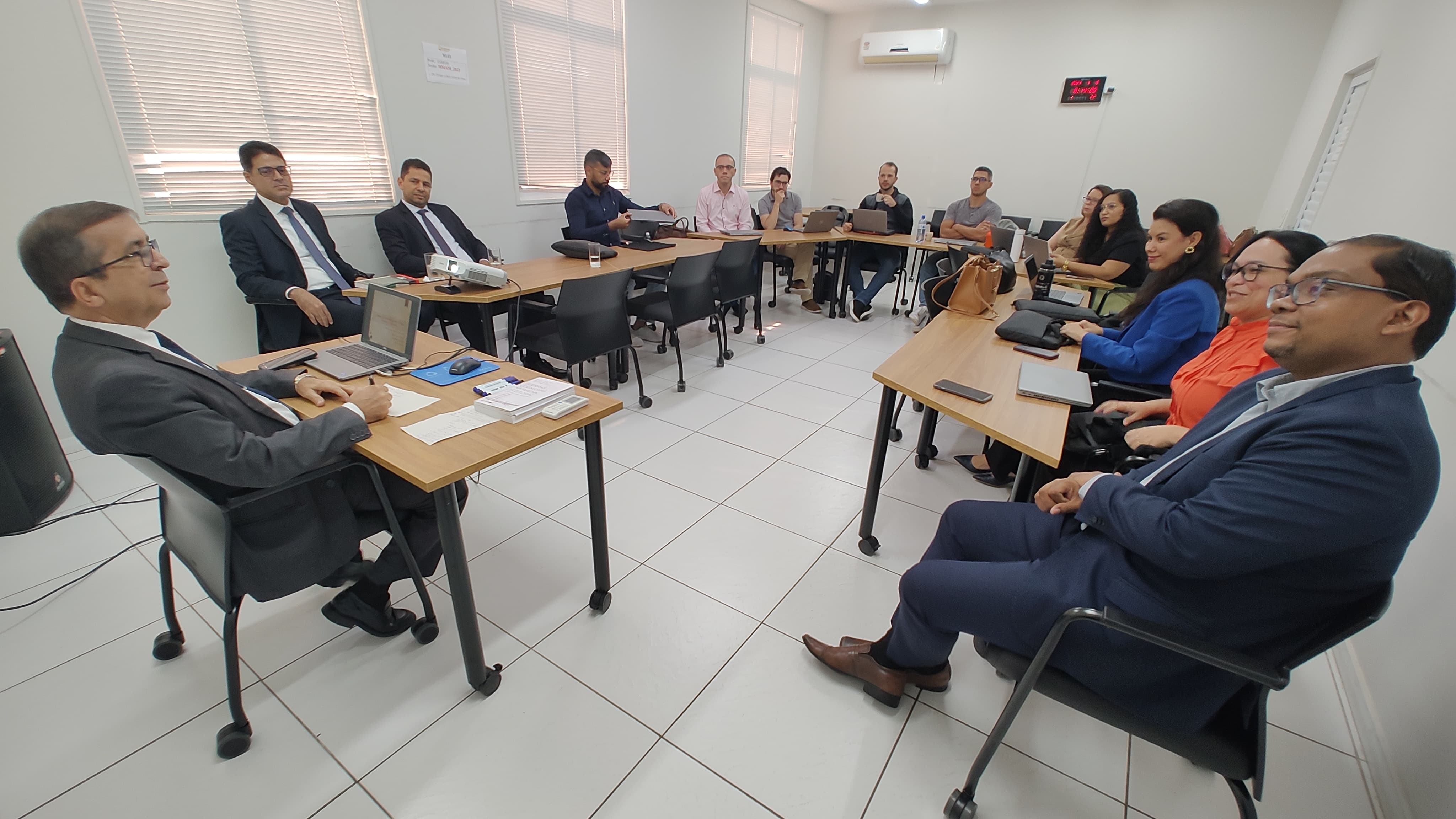 Fotografia horizontal. mostra sala de aula da esmam, que possui piso, teto e paredes brancas. alunas e alunos estão sentados e dispostos em formato circular e ouvem atentamente a exposição feita pelo desembargador josé gonçalo, presidente do tribunal regional eleitoral, que está à frente da sala.