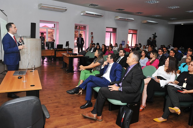 Foto colorida. Um homem de terno e gravata está em pé falando ao microfone, enquanto uma plateia está sentada em auditório assistindo à palestra.