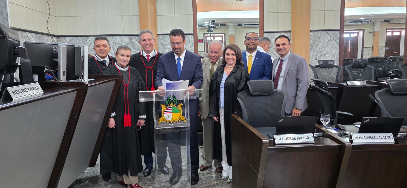 Fotografia horizontal, tirada no plenário do Tribunal de Justiça, que mostra oito pessoas posando para a câmera em torno de um púlpito de acrílico transparente com o brasão do Estado do Maranhão e a inscrição "Poder Judiciário". Ao centro, atrás do púlpito, um homem de terno azul-marinho, camisa branca, gravata azul e óculos segura algumas folhas de papel e sorri levemente.

À esquerda do homem central, três magistrados vestem togas pretas com medalhões vermelhos: um homem de cabelos escuros, uma mulher de cabelos grisalhos, com gola branca rendada, e um homem de cabelos grisalhos. À direita, um senhor de cabelos brancos e terno bege está ao lado de uma mulher de cabelos escuros, que veste toga preta sobre blusa clara. Logo atrás, um homem de óculos, terno azul e gravata amarela sorri. Na extremidade direita, um homem de terno cinza e gravata vinho também posa sorrindo. Ao fundo, é possível ver outra pessoa parcialmente encoberta.

O ambiente é o plenário, com paredes de mármore, colunas de madeira, bancadas de madeira escura, computadores e cadeiras pretas de espaldar alto. À esquerda, uma placa indica "Secretaria". À direita, as placas das bancadas trazem os nomes "Des. Jorge Rachid" e "Desa. Angela Salazar", que identificam os lugares dos desembargadores, e não as pessoas na foto.